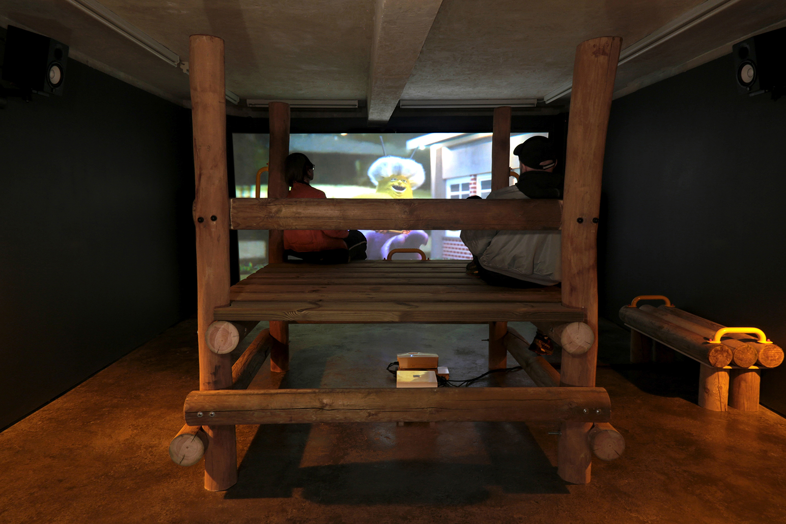 Two people sit on a large wooden climbing-frame-like structure in a dimly lit room, facing a projected animated film on the wall. The installation has a cozy, immersive feel, with speakers mounted on the walls and warm lighting accentuating the wood.
