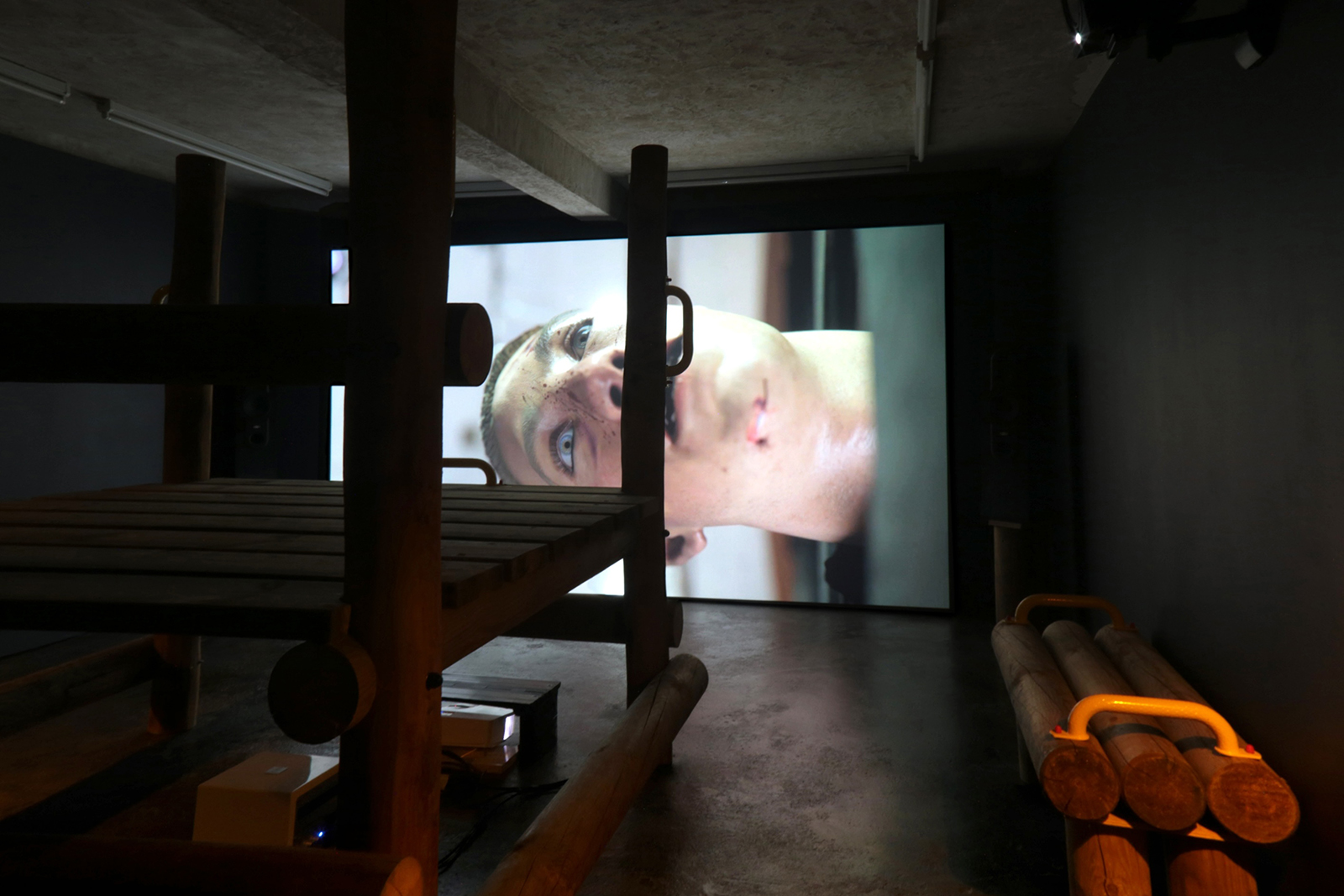Dark gallery space with wooden bench-like structures facing a large video projection showing a close-up of a person’s face.
