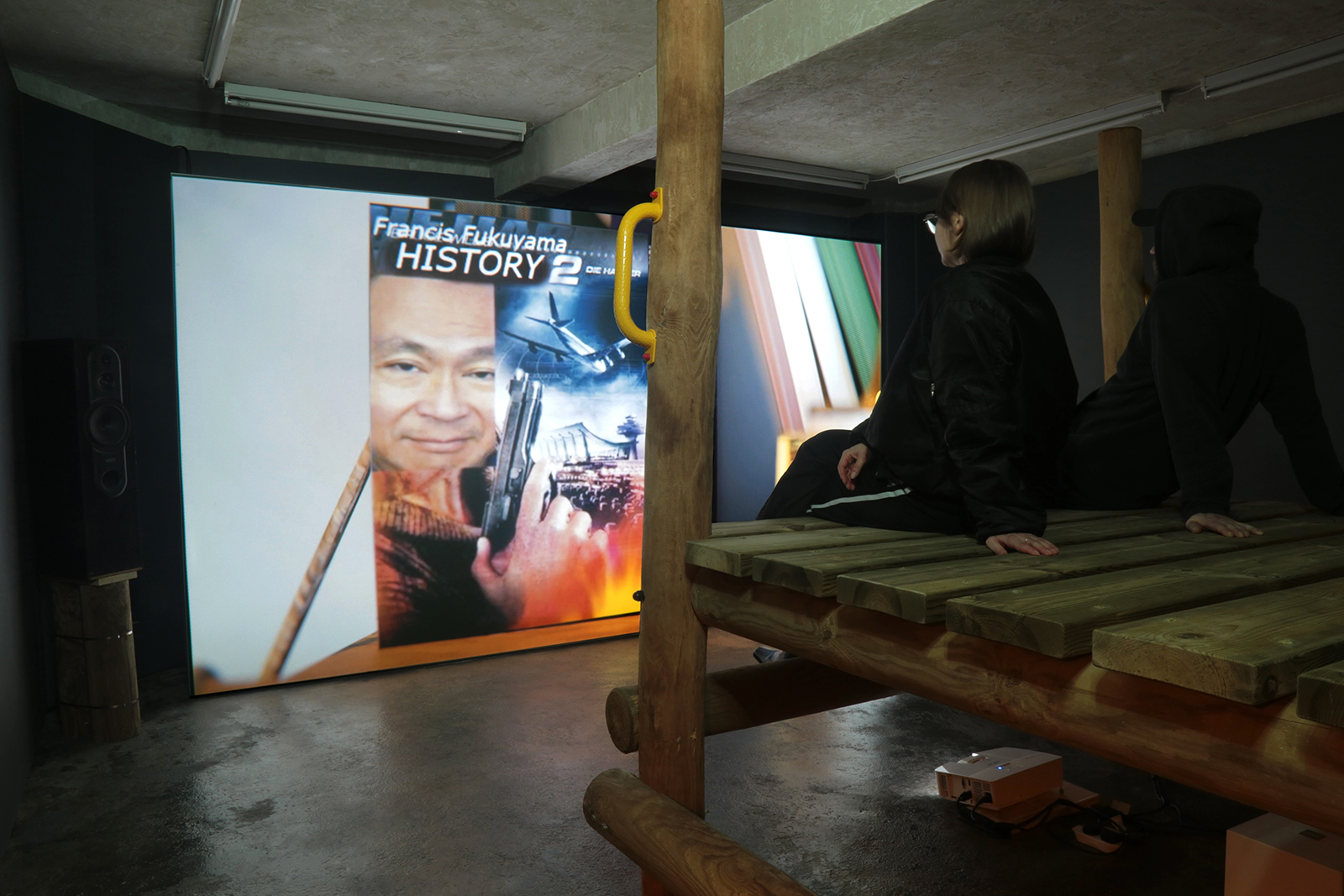 Two visitors sit on wooden platform seating in a dark gallery, watching a large video projection with colorful imagery and text.