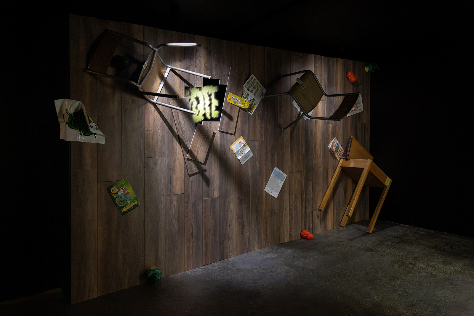 A darkly lit gallery, with a wall covered in brown floorboard. Different coloured objects and two chairs and attached to the wall, with a hole with floorboards missing, revealing a LED display which reads 'IDTE' in fuzzy green letters