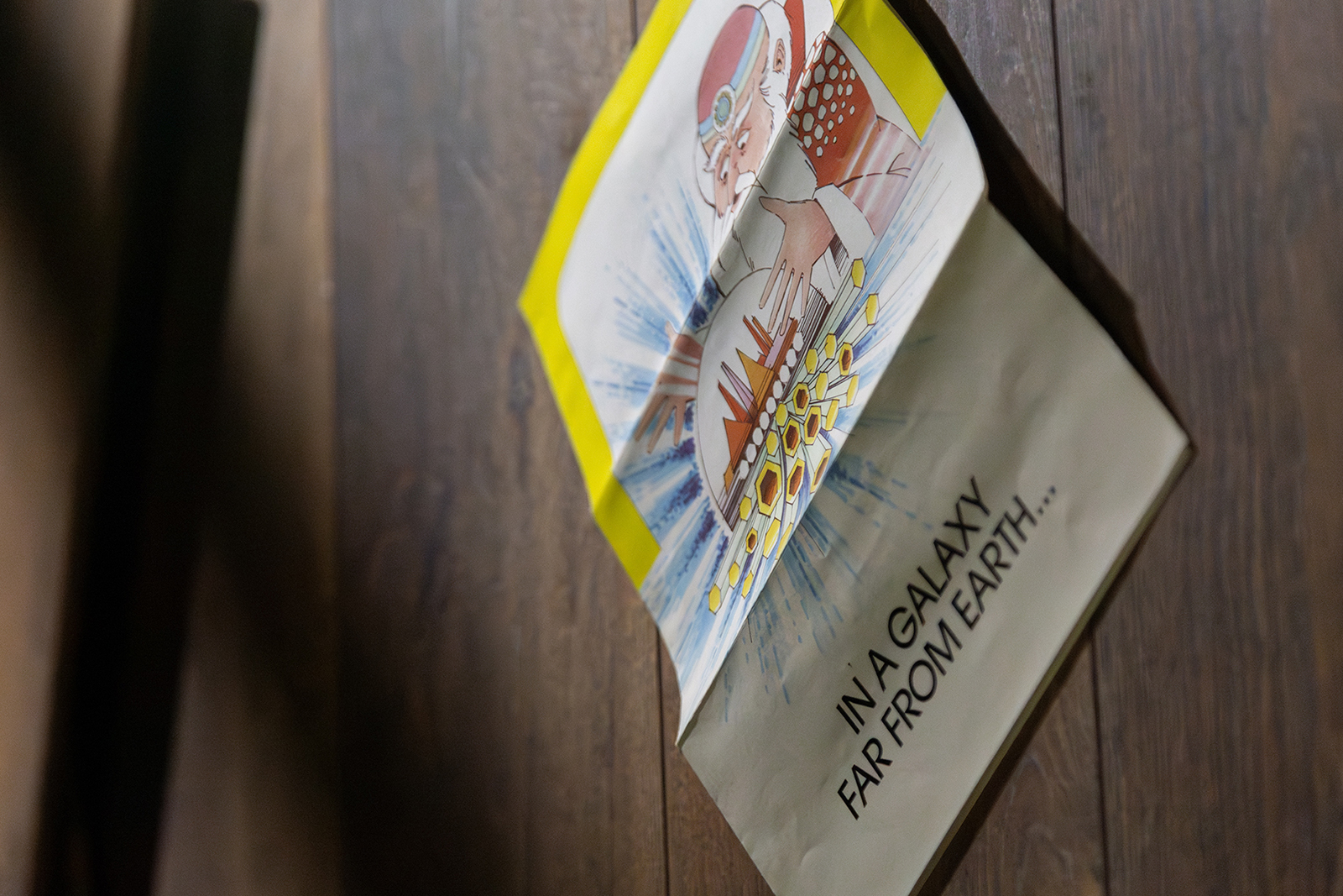 A piece of paper with a colourful cartoon of a wizard with his hands on either side of a crystal ball, inside which is a city with skyscrapers.  Words read underneath 'IN A GALAXY FAR FROM EARTH...', on background of brown floorboards