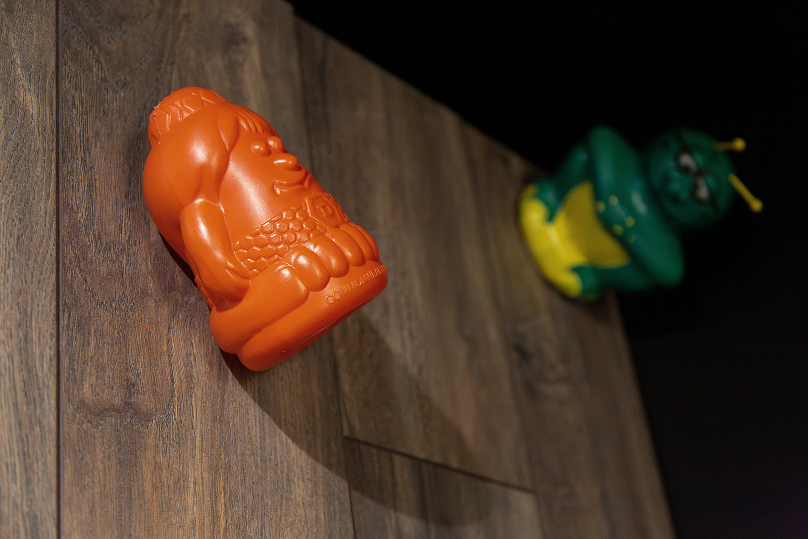 A orange plastic toy, of a smiling cartoon character with a wide body, attached to wooden floorboards.  A green alien-like creature toy sits in the background, slightly out of focus.