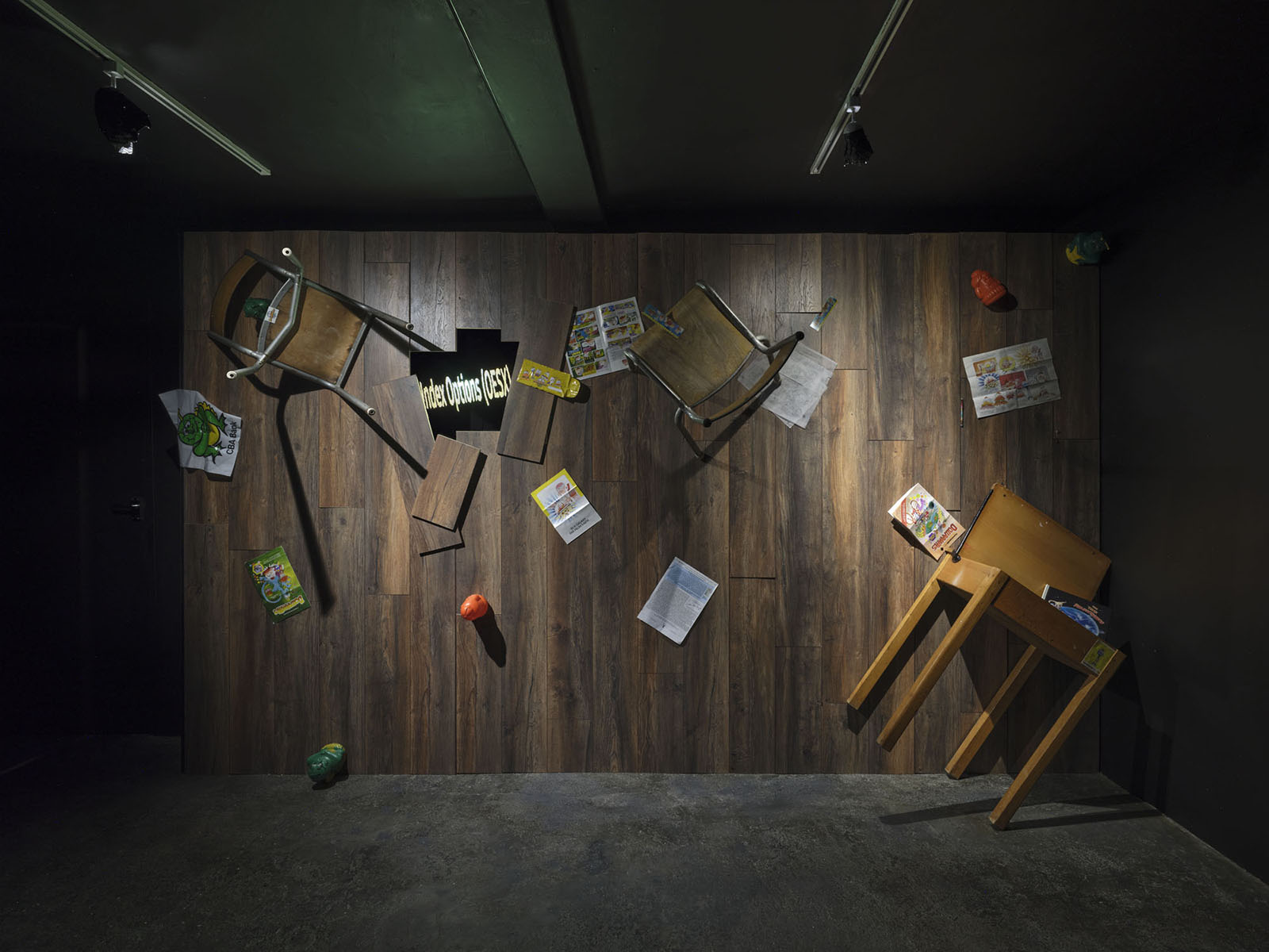 a view of of a darkly lit gallery with a wall covered in brown floorboard. Different coloured objects and two chairs and an old school desk attached to the wall, with a hole with floorboards missing, revealing a LED display which reads 'Index Options [ODSX]' in fuzzy green letters